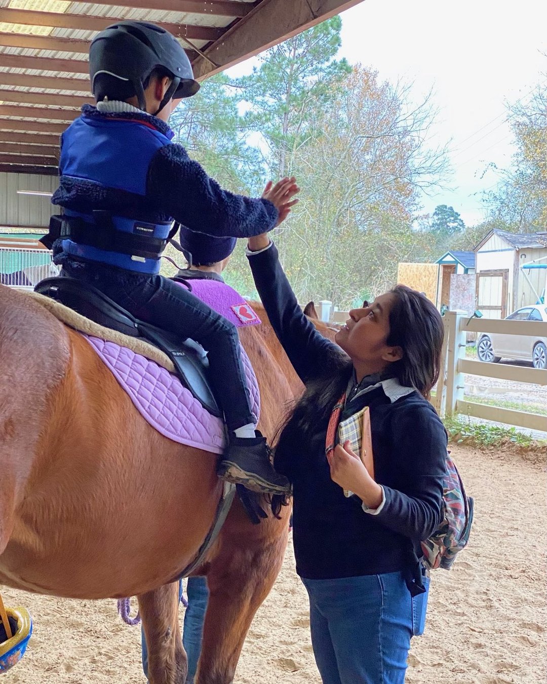 A young rider in a helmet and safety vest reaches down from the saddle for a high five.