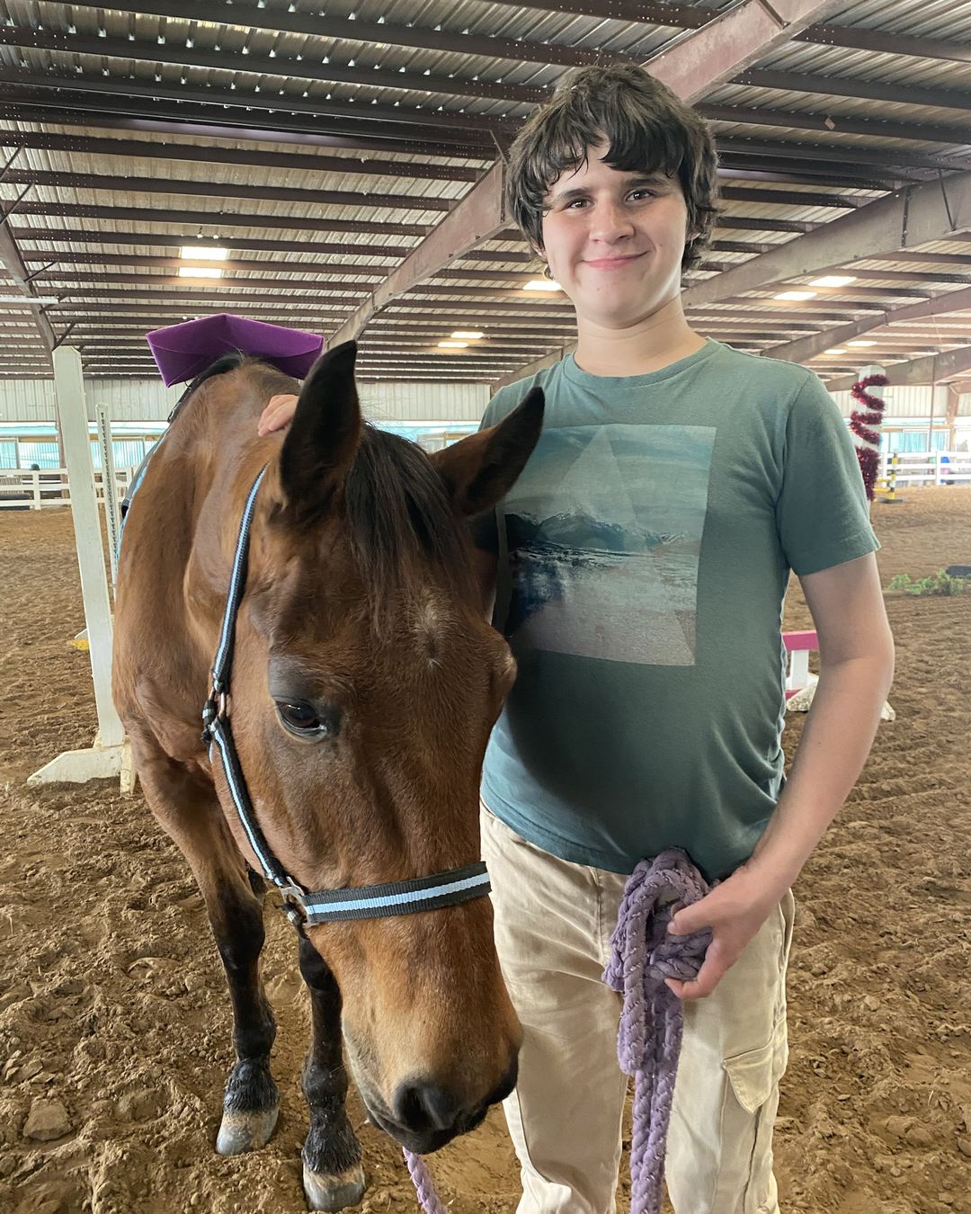 A smiling teen stands beside a bay horse in an indoor arena, holding the lead rope.